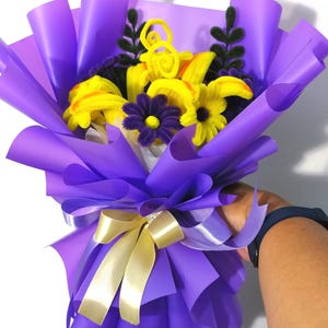 May include: A bouquet of handcrafted pipe cleaner flowers featuring bright yellow lilies and dark purple daisies with black centers. The arrangement includes dark green pipe cleaner foliage and is wrapped in vibrant purple paper with a pale yellow ribbon.