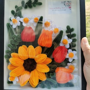 May include: A framed floral arrangement featuring a large yellow sunflower, orange tulips, and white daisies with orange centers. Green stems and leaves surround the flowers. The frame is black, and a small card with text is visible.