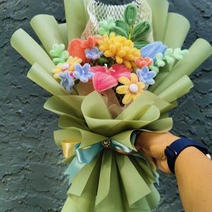 May include: A colorful bouquet of handmade felt flowers, including yellow, blue, orange, and pink blooms, wrapped in light green paper. The bouquet is tied with a light blue ribbon and a gold ribbon, creating a cheerful and artistic floral arrangement.