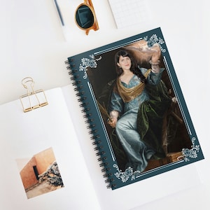 以下が含まれることがあります： 白い縁取りのある青いスパイラルノート。青い緑のドレスを着た女性が白い鳥を持っている絵が描かれている。絵は白いサイコロのデザインが施された装飾的な縁取りで囲まれている。