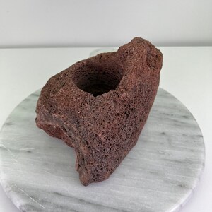 May include: A reddish-brown, porous lava rock with a central opening, possibly for a candle or small plant. The rock sits on a white and gray marble-patterned surface, showcasing its natural texture and color.