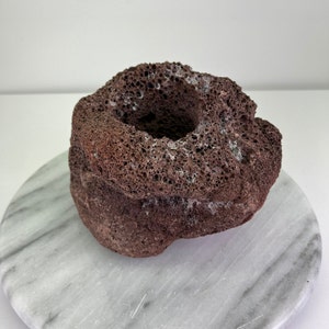 May include: A porous, dark reddish-brown lava rock candle holder. The rock has a cylindrical shape with a central cavity for a candle. The surface is textured with numerous small holes. It sits on a round, grey and white marble surface.