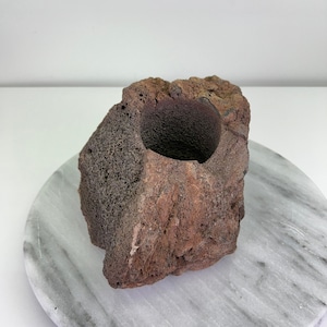 May include: A natural, rough-hewn rock with a large, circular opening. The rock is a mix of reddish-brown and dark grey, with a porous texture. The opening reveals a darker, smoother interior. The object is likely a decorative item or a unique container.