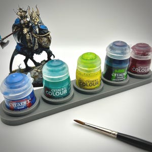 May include: A gray paint holder with five Citadel Colour paint pots in blue, green, yellow, blue-green, and red. A paintbrush lies to the right. A miniature figure of a warrior on a beast is in the background.