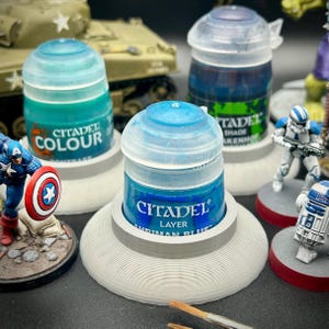 May include: Three Citadel paint pots in plastic holders. The front pot is labeled Citadel Layer Ahriman Blue. To the left is a turquoise Contrast paint pot, and to the right is a dark blue Shade paint pot. Miniature figures and a tank model are visible.