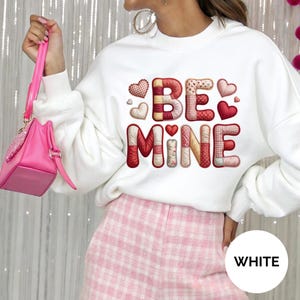 Może przedstawiać: Biała bluza z napisem "BE MINE" w patchworkowym wzorze z sercami. Litery i serca są w odcieniach czerwieni, różu i kremu. Top z motywem walentynkowym.
