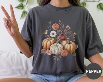 May include: Dark grey t-shirt with a watercolour design of pumpkins, flowers, and a small bird. The pumpkins are orange and white, with flowers in shades of orange, pink, and white. The shirt has a relaxed fit.