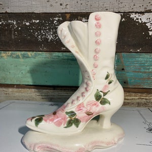 May include: A vintage, off-white ceramic boot-shaped vase. The boot features pink floral designs, pink button accents, and a decorative base. The heel is a small, curved design. The vase is a unique decorative item.