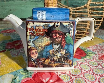 Może przedstawiać: Biały ceramiczny czajnik z niebiesko-brązową ilustracją pirata, mapą skarbów i napisem "Treasure Island". Czajnik ma pokrywkę w kształcie skrzyni skarbów.