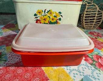 May include: A red plastic storage container with a white lid. The lid has a slightly rounded shape and a smooth surface. The container is sitting on a colorful patterned fabric.
