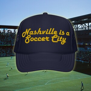 May include: A navy blue trucker hat with a yellow embroidered design that says "Nashville is a Soccer City".