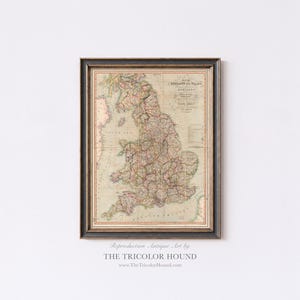 May include: Framed antique map of England and Wales, featuring detailed cartography in muted tones of beige, pink, and green. The map is enclosed in a black and gold frame. Text at the bottom reads "Reproduction Antique Art by THE TRICOLOR HOUND".