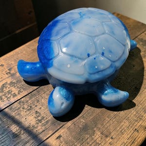 May include: A turtle-shaped decorative object crafted from resin with a glossy finish. The shell and limbs feature a marbled pattern of deep royal blue and opaque white, creating a swirling effect across the smooth, rounded surface of the figure.