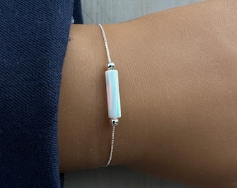 May include: A delicate silver bracelet featuring a rectangular, iridescent opal-like bead. The bead is centered on a fine silver chain, with a small silver bead on each side. The bracelet is shown on a wrist, with a navy blue sleeve in the background.