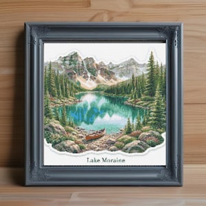 Può includere: Un ricamo a punto croce incorniciato raffigurante un paesaggio del lago Moraine con acqua turchese, una canoa rossa sulla riva, alberi sempreverdi e montagne innevate. Il testo Lake Moraine è stampato in nero nella parte inferiore dell'opera.