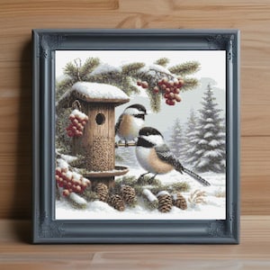Könnte beinhalten: Gerahmtes Kreuzstich-Kunstwerk mit zwei Kohlmeisen, die sich in einer verschneiten Winterszene in der Nähe eines hölzernen Vogelhauses aufhalten. Das Kunstwerk zeigt Tannenzapfen, rote Beeren und immergrüne Zweige, alles vor dem Hintergrund schneebedeckter Bäume. Der Rahmen ist grau.