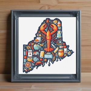 May include: Framed cross-stitch art depicting the state of Maine, filled with pixelated images of local icons. A large orange lobster dominates the center, surrounded by food, landmarks, and the word "MAINE". The frame is a dark gray color.