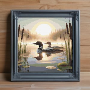 May include: Framed cross-stitch artwork depicting two loons on a lake at sunrise. The scene includes cattails, lily pads, and a hazy sun. The frame is a dark gray with ornate detailing.