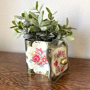 May include: A vintage floral tin container with a lid, featuring a gold trim and a decorative floral design. The container holds a small green plant. The lid is partially open, revealing the interior. The container is on a wooden surface.