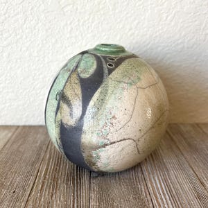 May include: A spherical ceramic vase featuring a textured, crackled off-white surface with mottled green and black abstract patterns. The vessel has a small, circular opening at the top with a smooth green rim and dark decorative markings.