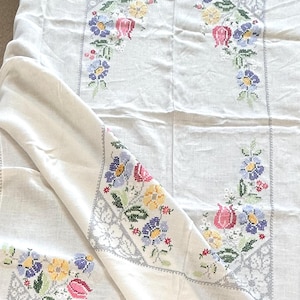 May include: White tablecloth with embroidered floral designs in pink, yellow, blue, and green. The embroidery is framed by a gray geometric border. The cloth is folded, showing the detailed needlework.