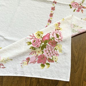 May include: White tablecloth with a floral design featuring pink roses, pears, grapes, and yellow accents. The tablecloth has a detailed border with similar floral patterns. The fabric is folded, revealing the pattern.