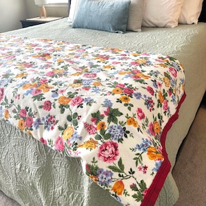 May include: A bed with a floral patterned blanket. The blanket features a white background with a colorful array of flowers in shades of pink, yellow, orange, and blue. The blanket has a red border and is draped over a bed with green bedding and pillows.