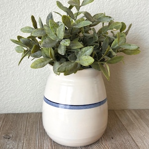 May include: A white ceramic vase with a blue stripe, filled with green leafy faux foliage. The vase has a rounded shape and sits on a wooden surface. The leaves have a dusty green appearance.
