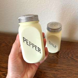 May include: A pair of vintage salt and pepper shakers. The shakers are a pale yellow color with silver metal lids. The pepper shaker has the word "PEPPER" printed in black, and the salt shaker has the word "SALT" printed in black.