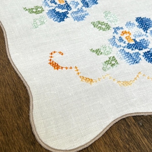 May include: White linen table runner with embroidered blue floral design. The flowers have orange centers and green leaves. The runner has a scalloped edge with a tan border. The embroidery is detailed and the fabric appears to be linen.
