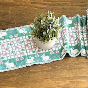 May include: A green and white table runner with a repeating pattern of bunnies and flowers. The runner has a frayed edge and is perfect for a spring or Easter table setting.