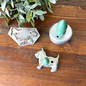 May include: Three vintage metal cookie cutters on a wooden surface. One is a frog shape, another is a round cutter with a green handle, and the third is a dog shape with a green accent. Green foliage is in the background.