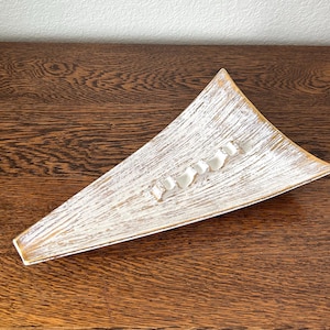 May include: Triangular ashtray with a white and gold finish. The ashtray has a textured surface and four small rests for cigarettes. The ashtray is made of a light-colored material and sits on a wooden surface.