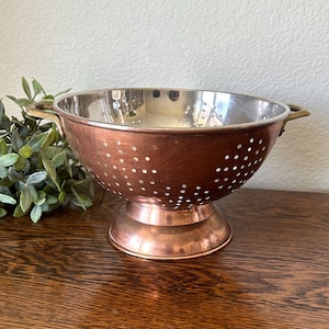 May include: A round copper colander with a polished silver-toned interior, featuring a wide pedestal base and two small side handles. The exterior surface is made of warm-toned copper and is perforated with numerous small circular drainage holes.