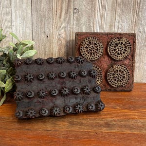 May include: Two rectangular wooden printing blocks with carved designs. One block features rows of small star and moon motifs in dark brown wood. The second block is reddish-brown with four circular patterns containing intricate floral-like cutouts.