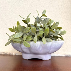 May include: A light purple ceramic planter with a scalloped rim, filled with green leafy faux plants. The planter sits on a small pedestal base. The leaves have a dusty, textured appearance. The planter is on a dark brown surface.