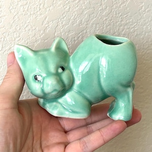 May include: A mint green ceramic cat figurine with black eyes. The cat is in a playful pose, with its front paws extended and a small opening on its back. This decorative item is a unique addition to any home decor.