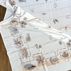 May include: White tablecloth with a repeating pattern of leaves in shades of brown, gray, and beige. The leaves are framed in square and rectangular shapes with gold-colored lines. The tablecloth is folded, showing the pattern.
