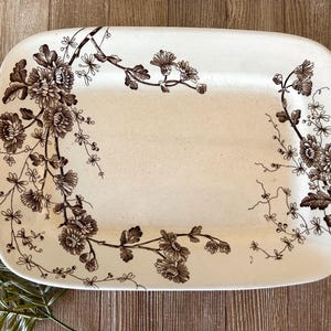 May include: A rectangular ceramic serving platter with a cream-colored base and a dark brown floral transferware pattern. The design features detailed illustrations of chrysanthemum-like flowers and delicate vines extending along the edges.