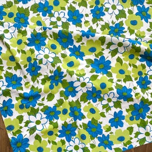 May include: A white fabric featuring a repeating floral pattern with bright blue and lime green flowers, along with green leaves. The textile displays a dense arrangement of blossoms in varying sizes, creating a continuous botanical design across the surface.