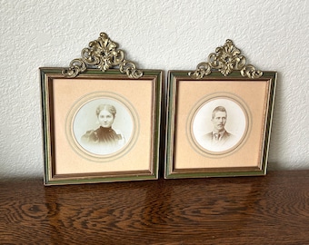 May include: Two antique framed portraits. Each portrait features a sepia-toned image within a circular frame, set against a light peach background. The frames are green with gold filigree accents at the top. The portraits are displayed on a wooden surface.