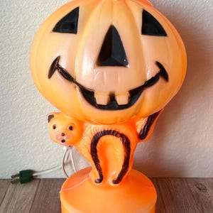 May include: A vintage orange plastic blow mold lamp featuring a smiling jack-o'-lantern head with black triangular eyes and a nose, and two square teeth. A small orange cat figure is attached to the side, and the lamp has a white power cord.