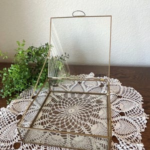 May include: A clear glass and brass display box with a hinged lid. The box sits on a white lace doily. The interior of the box features a white lace pattern. A small brass handle is on the lid.