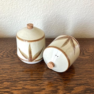 May include: A pair of ceramic jars with cork stoppers. The jars are off-white with brown leaf-like designs and a brown rim. One jar is upright with a lid, the other is on its side. The jars are on a wooden surface.