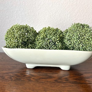 May include: A white ceramic planter with three spherical, green, textured plant arrangements. The planter is oval-shaped and rests on three small feet. The arrangement sits on a dark wooden surface against a white wall.