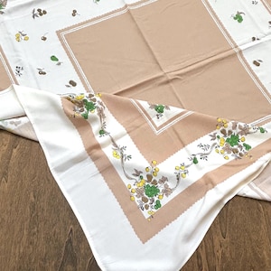 May include: A vintage tablecloth with a white and tan color scheme. The tablecloth features a repeating pattern of green, yellow, and brown fruit and floral designs. The edges have a scalloped border.