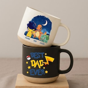 May include: Two ceramic mugs stacked on a wooden surface. The top mug is off-white with a cartoon illustration of Pikachu, Charmander, and Bulbasaur. The bottom mug is black with the text "BEST DAD EVER" and a lightning bolt design.