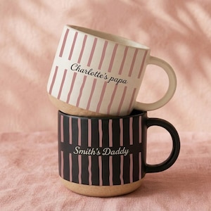 May include: Two stacked ceramic mugs. The top mug is white with pink vertical stripes and the text "Charlotte's papa". The bottom mug is black with pink vertical stripes and the text "Smith's Daddy". Both mugs have a tan base and a handle.