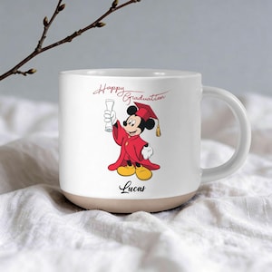 May include: White ceramic mug with a tan base, featuring a graduation-themed Mickey Mouse illustration. The character is dressed in a red graduation gown and cap, holding a diploma. The words "Happy Graduation" and the name "Lucas" are also printed on the mug.