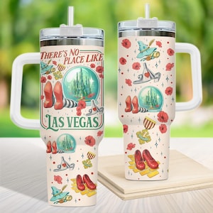 Puede incluir: Un vaso de viaje color crema con asa y tapa blancas, con ilustraciones del tema El Mago de Oz. El vaso tiene el texto "There's No Place Like Las Vegas" e imágenes de zapatillas rojas, un sombrero de bruja y una bola de cristal.